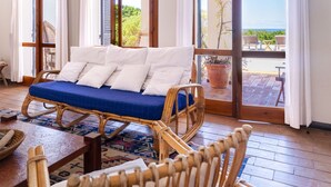 Un sofá de mimbre con cojines blancos y uno azul, ubicado en una habitación con ventanas amplias y vista al mar.
