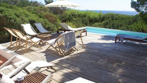 Terraza de madera con sillones de descanso y una pequeña mesa, con vista a una piscina y al mar.