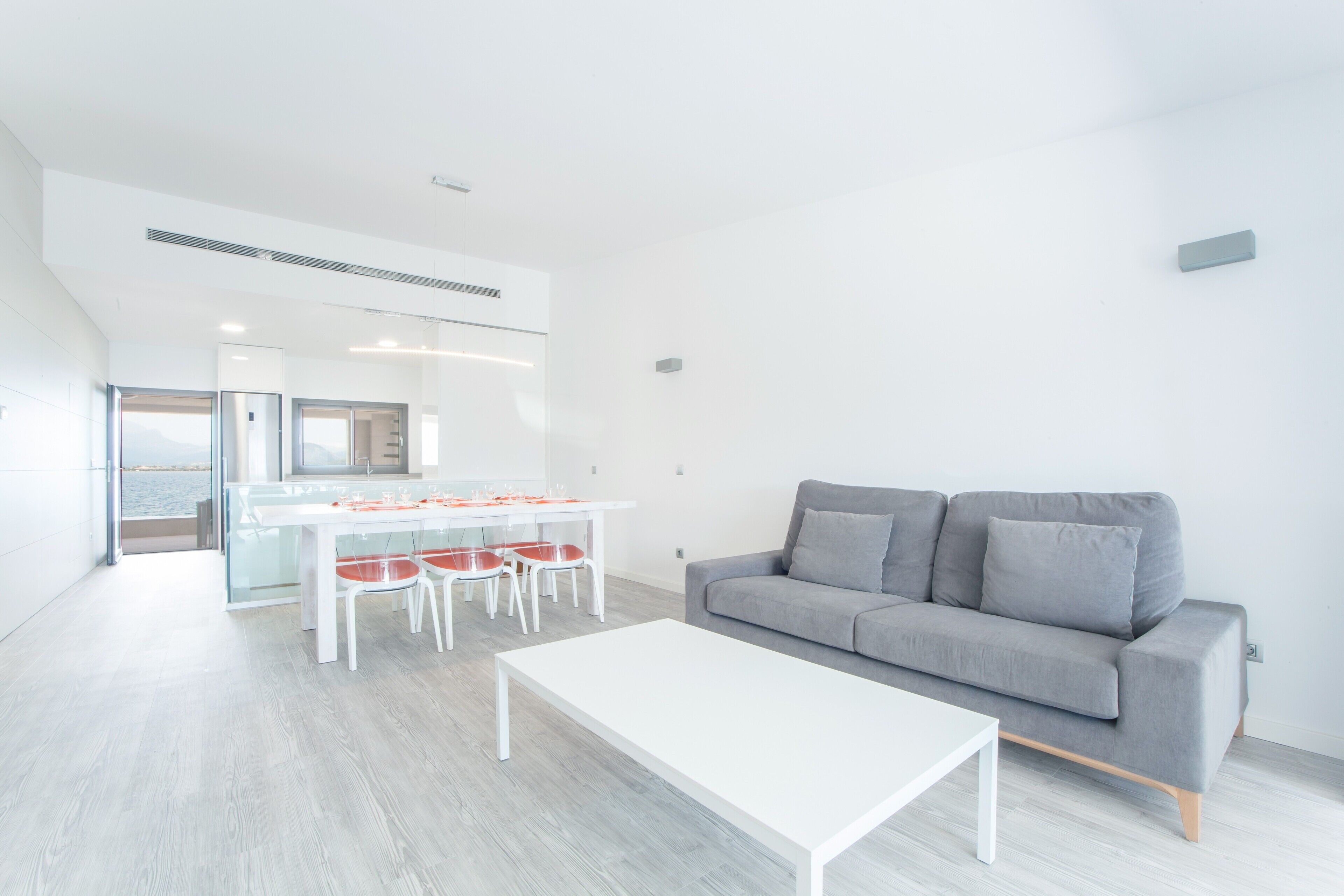 Una sala moderna con un sofá gris, una mesa de centro blanca y un comedor con una mesa blanca y sillas rojas.