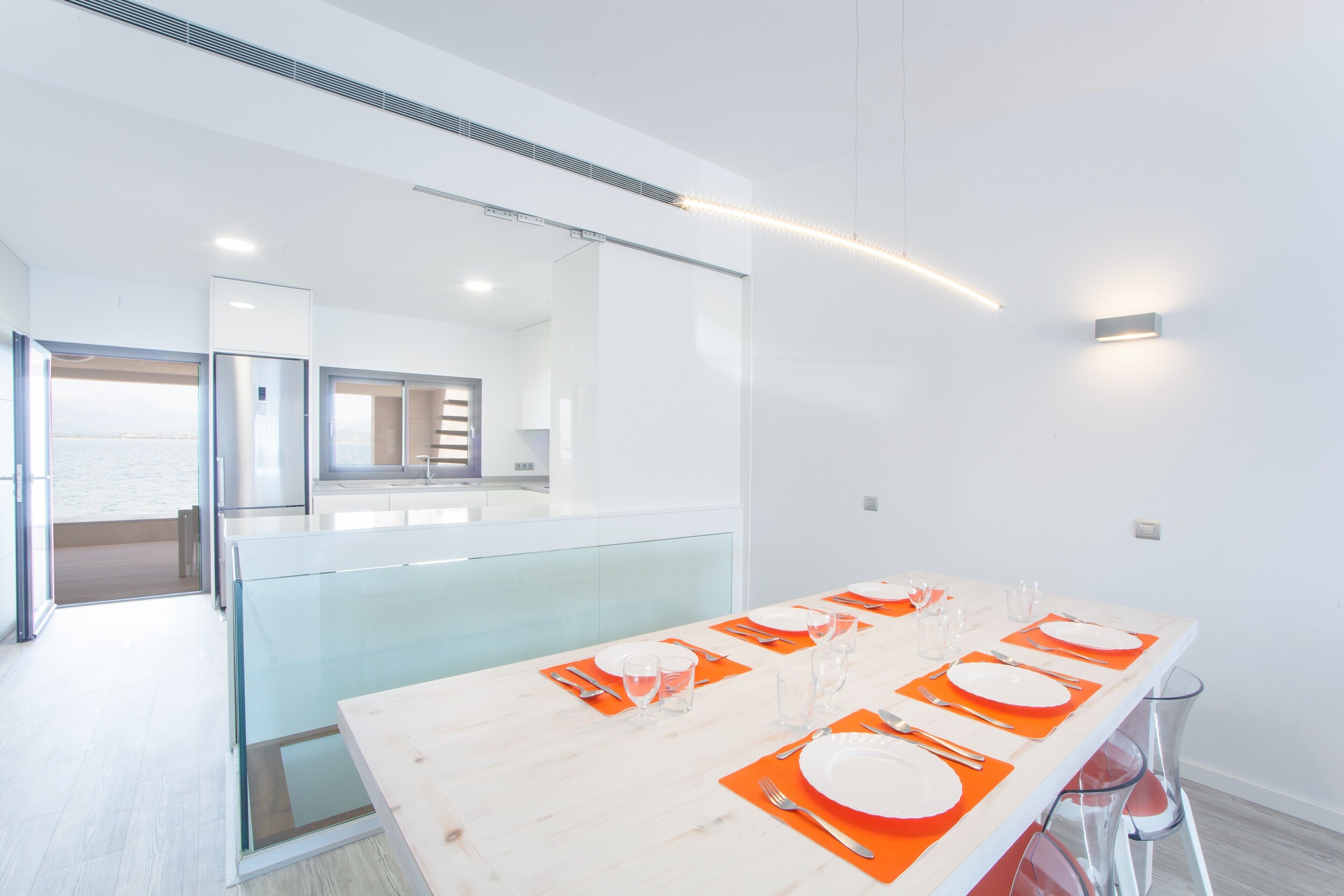 Un comedor moderno con una larga mesa dispuesta para seis personas, barandales de vidrio y vista a la cocina.