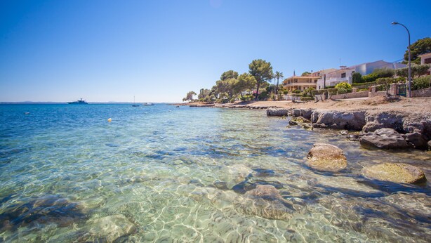Kystlandskab med klart vand, klipper og bygninger langs stranden.