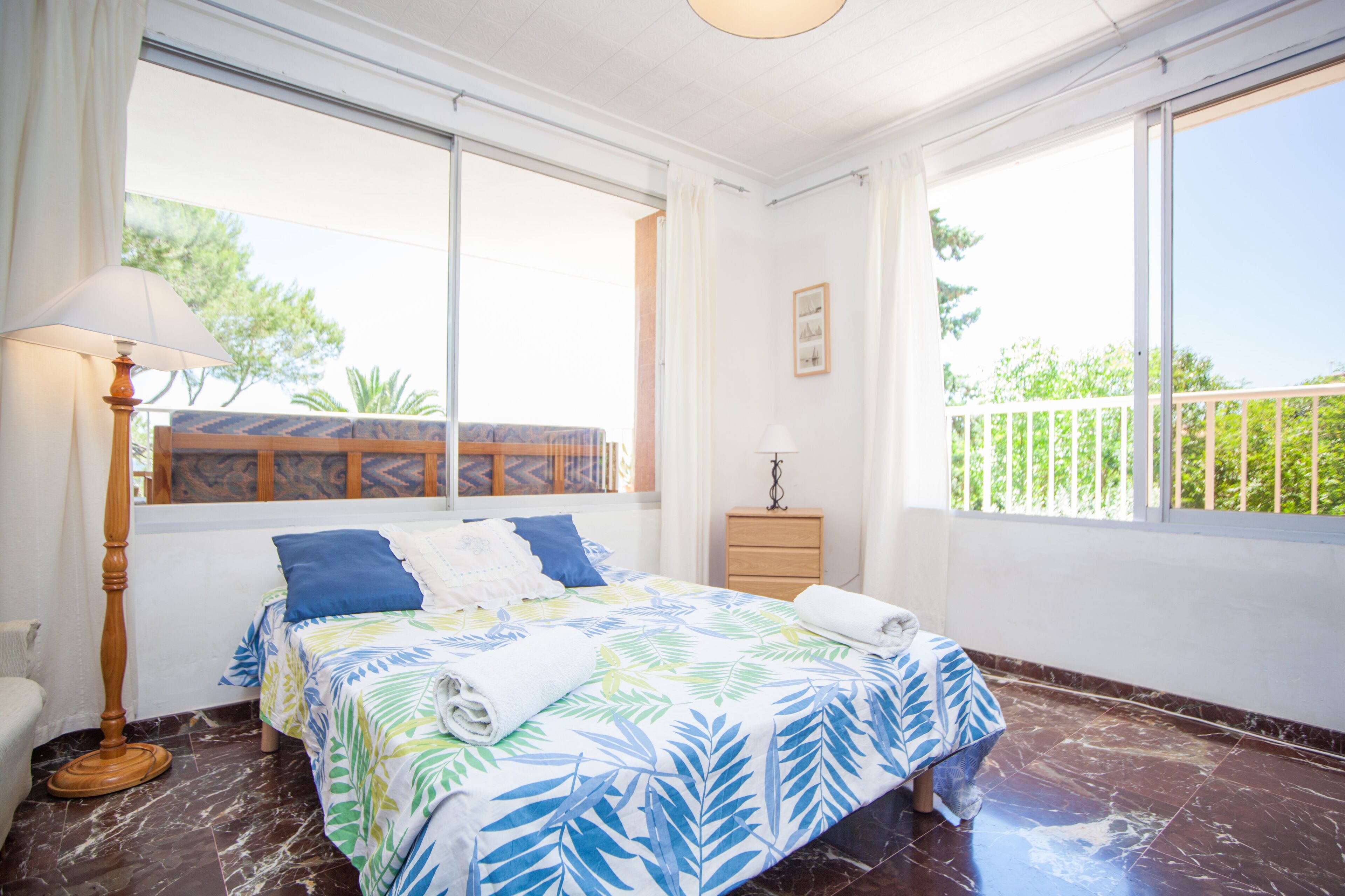 A bedroom with a bed, a nightstand, a lamp, and a window with a view of greenery.