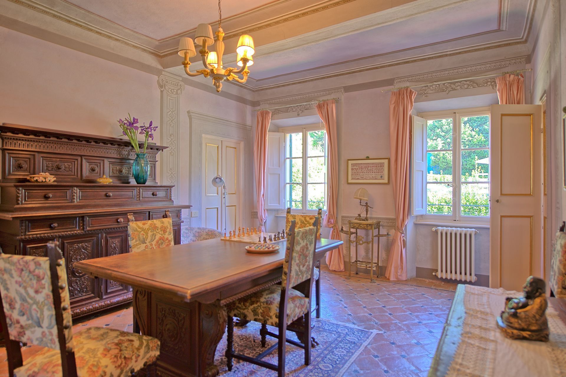 Ein Esszimmer mit Holztisch, floralen Stühlen, einer Kristallleuchter und Blick auf den Garten durch große Fenster.