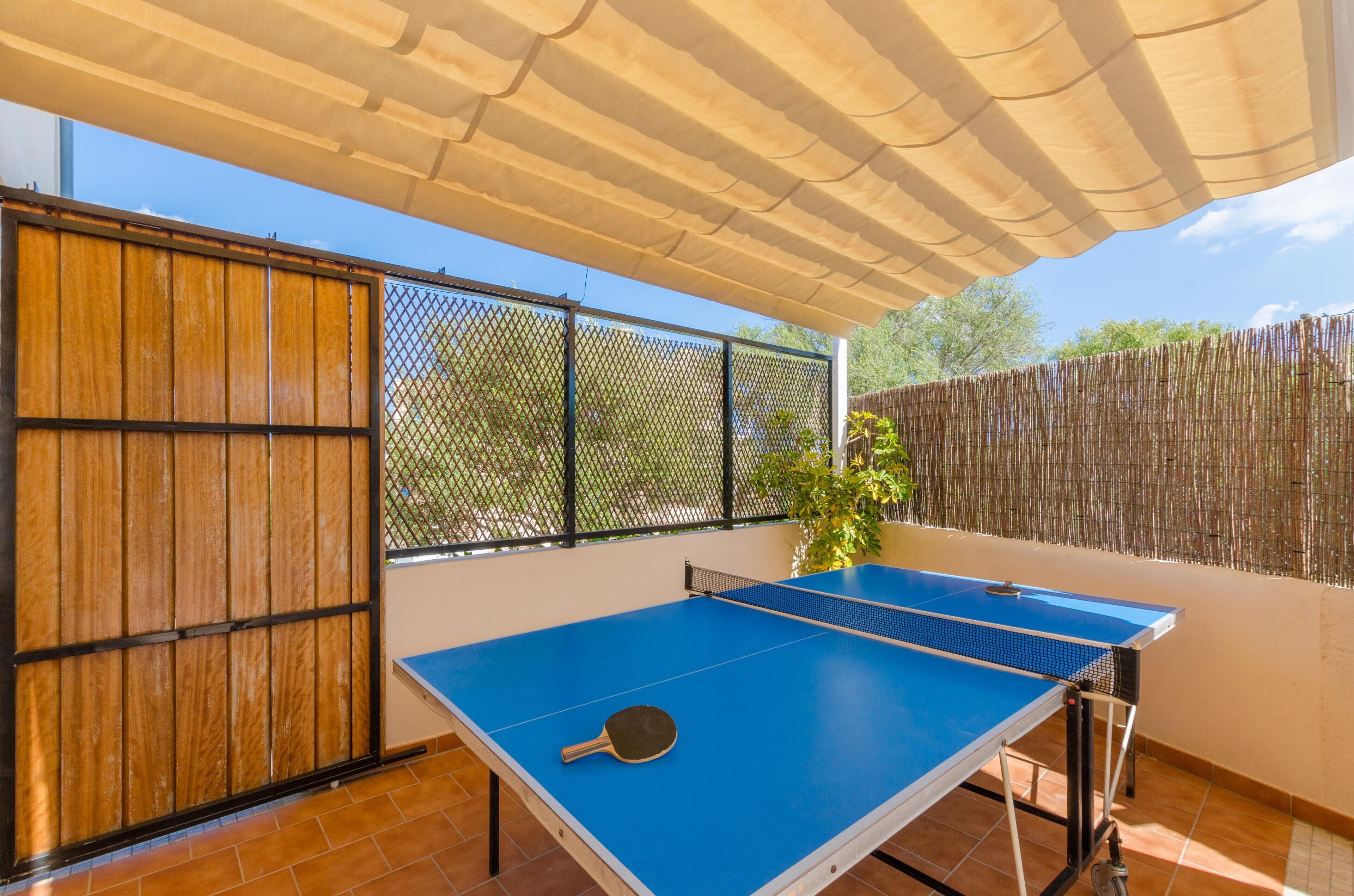 A ping pong table with a paddle on a sunny patio with a wooden fence and a mesh screen.