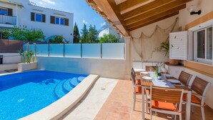 Un espace aménagé au bord de la piscine, avec une table et des chaises, une pergola et une vue sur un bâtiment doté de fenêtres et de plantes.