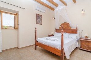 A bedroom with a wooden four-poster bed, a bedside table with a lamp, a nightstand, a window with a view, and a wall-mounted air conditioner.