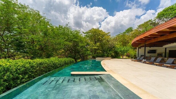 A swimming pool with a wooden deck and lounge chairs, surrounded by lush greenery.
