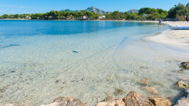 Une plage avec une eau claire et peu profonde, un rivage sablonneux et une montagne en arrière-plan.