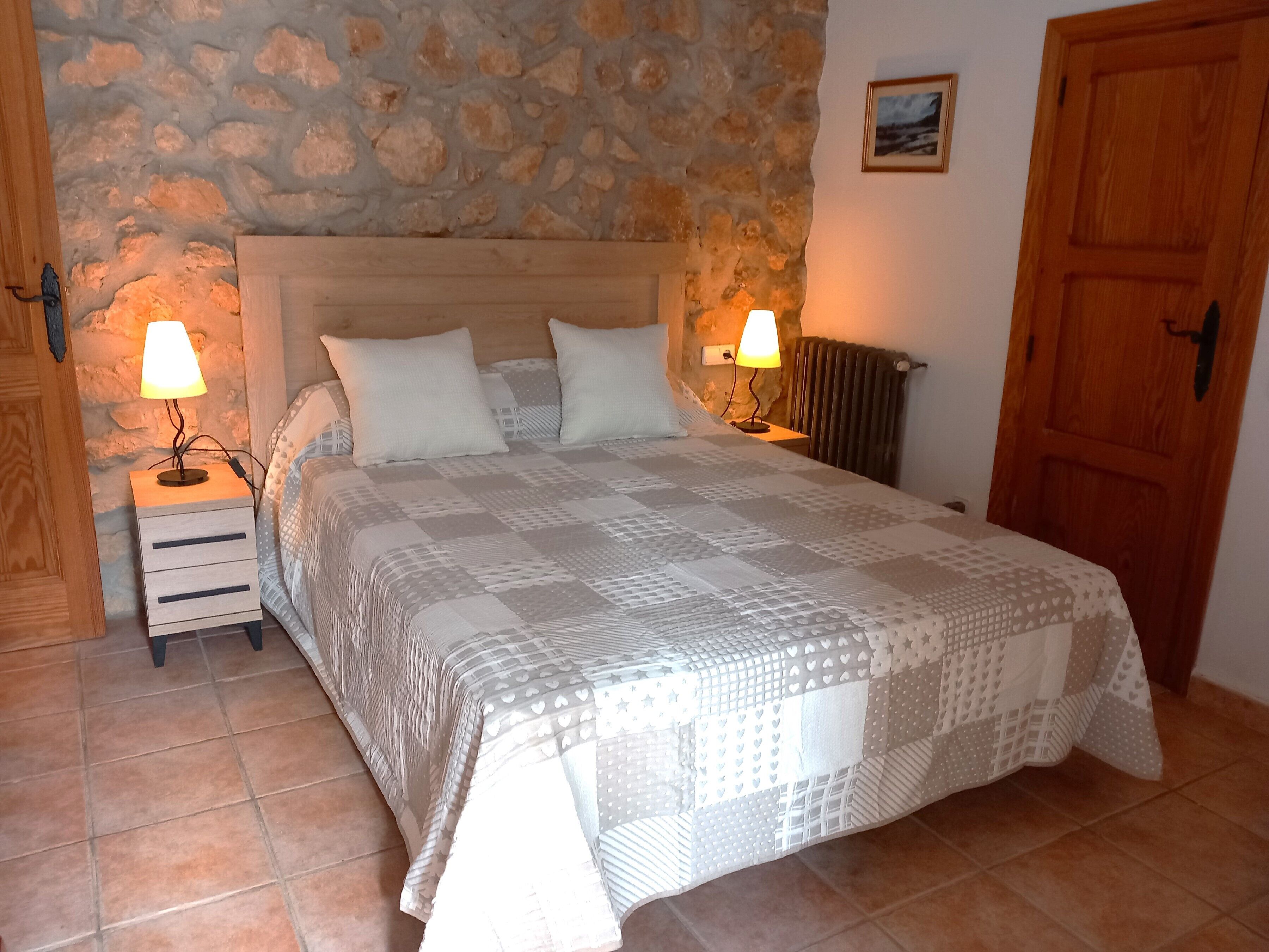 A bedroom with a stone wall, a wooden door, a bed with a patterned bedspread, two bedside lamps, and a small nightstand.