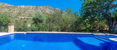 A clear blue swimming pool with a stone structure on one side, surrounded by trees and a mountainous backdrop.