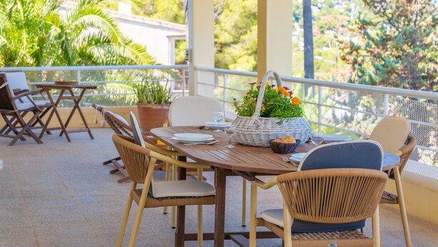 Um pátio externo com mesa de madeira preparada para refeições, cadeiras de vime e um cesto de vime com flores.