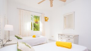 Um quarto claro e arejado, com duas camas, um ventilador de teto, uma janela com cortinas, um espelho e uma cômoda branca.