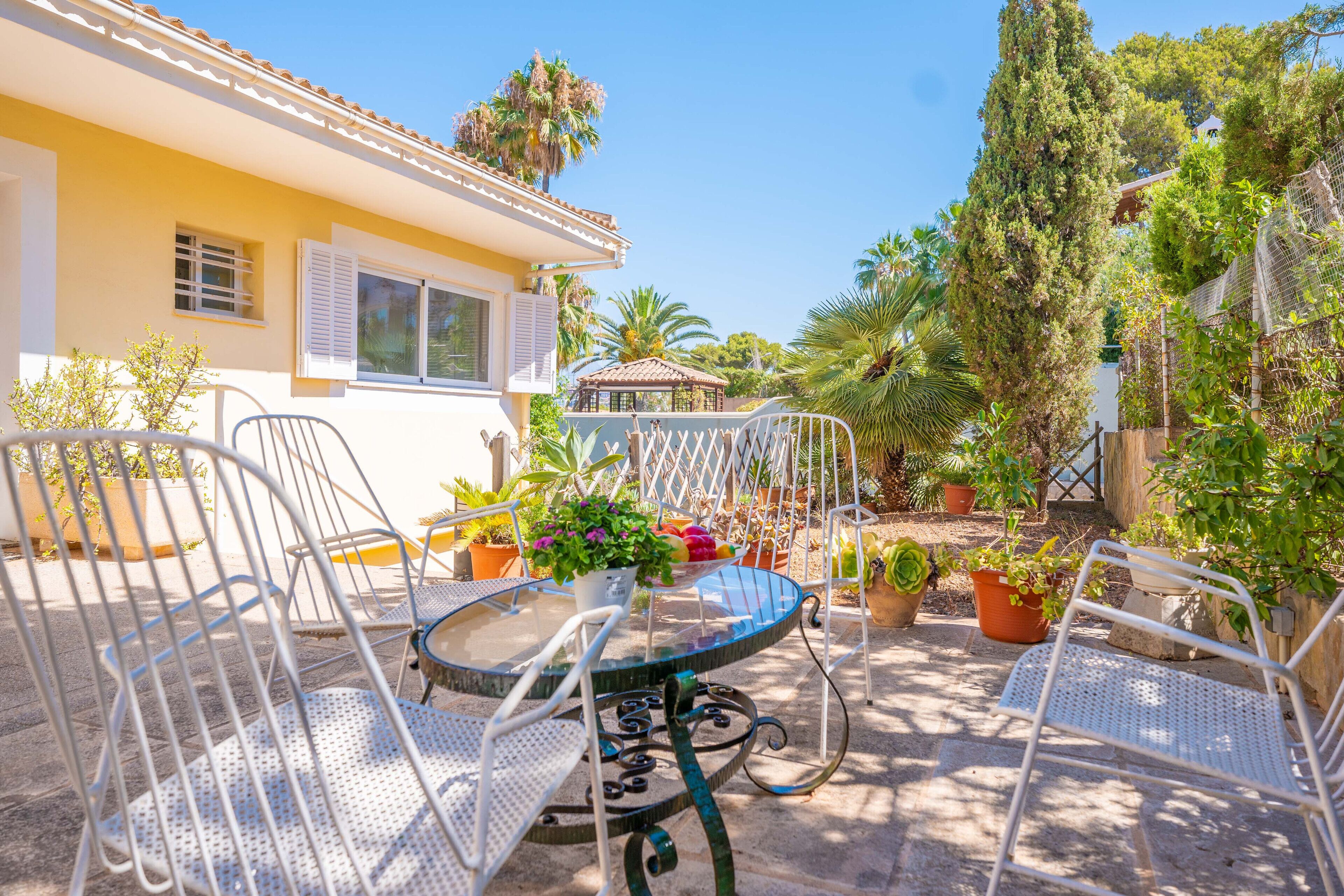 A patio with a round table and white metal chairs, surrounded by potted plants and a yellow building with shutters.