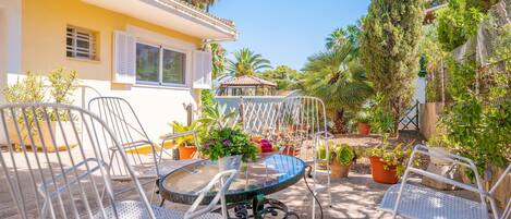 A patio with a round table and white metal chairs, surrounded by potted plants and a yellow building with shutters.
