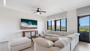 Una sala de estar moderna con un sofá modular, un pequeño otoman, una mesa de centro y un ventilador de techo. Hay un cuadro grande de una ola en la pared y vistas a un paisaje a través de amplias ventanas.