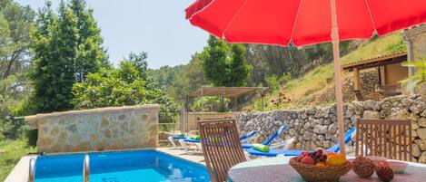 A poolside area with a stone wall, lounge chairs, and a table with a fruit basket.