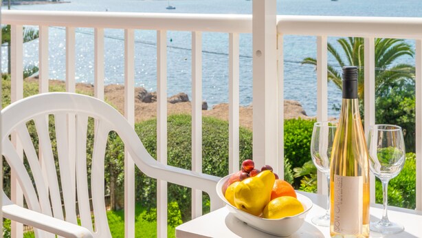 Una silla blanca, un cuenco de fruta, una botella de vino y dos copas de vino sobre una mesa con vistas al mar y las montañas.