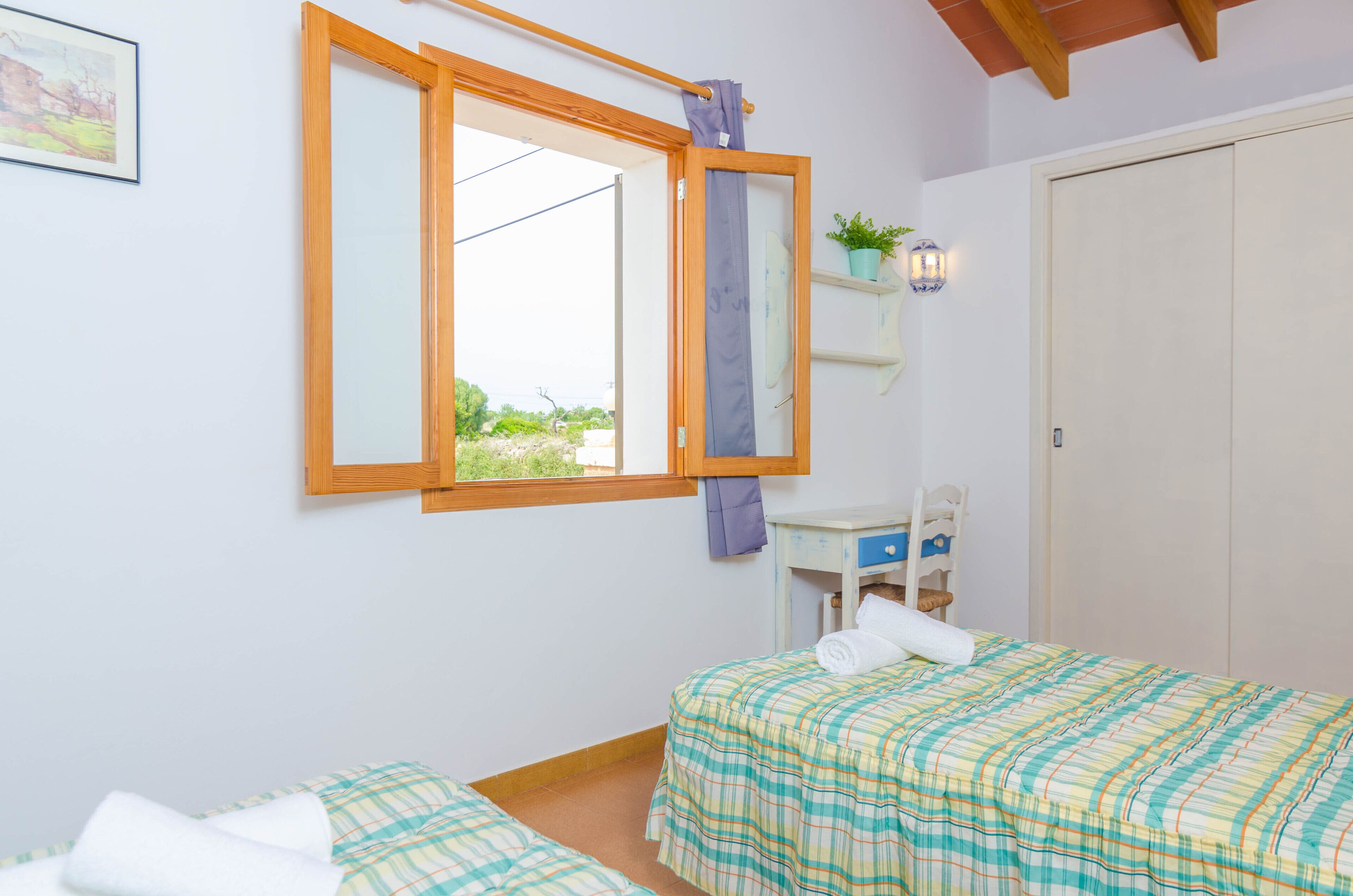A bedroom with two beds, a wooden-framed window, a small desk with a chair, a potted plant, and a door.