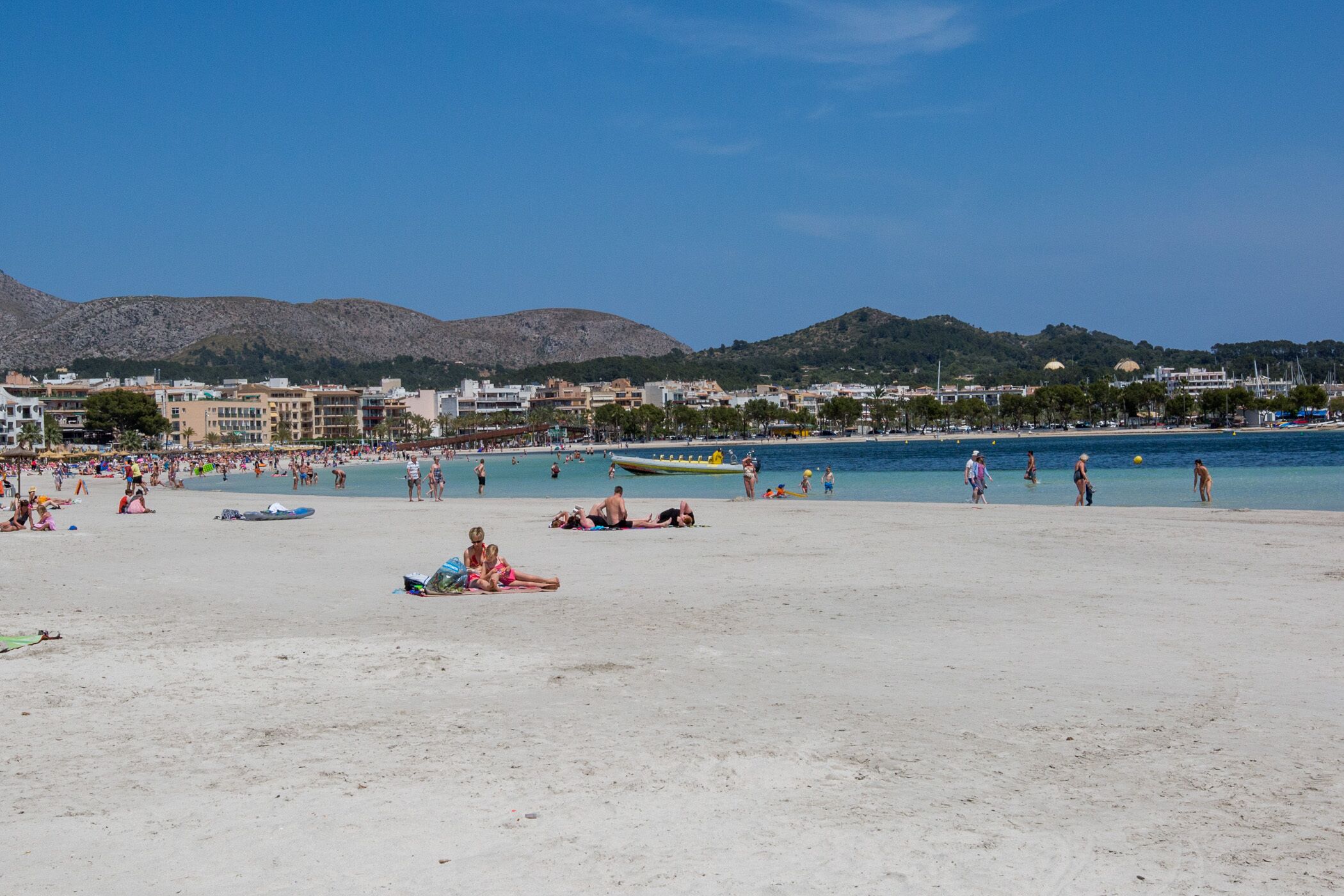 Una spiaggia con persone che prendono il sole e nuotano, barche e edifici sullo sfondo.