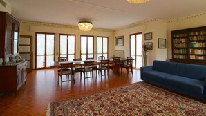 Un salón amplio con zona de comedor, ventanas grandes, una araña y un sofá azul, además de una estantería.