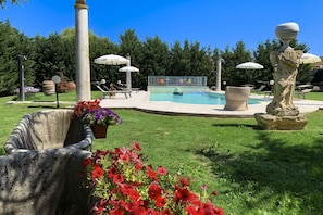 A swimming pool with lounge chairs and umbrellas, surrounded by greenery and a statue.