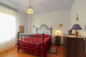 A bedroom with a bed, a chandelier, a desk with a lamp, and a small rug.