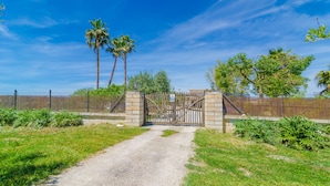 Une entrée fermée par un portail, avec un chemin de gravier menant à un bâtiment, le tout entouré de verdure et de palmiers.