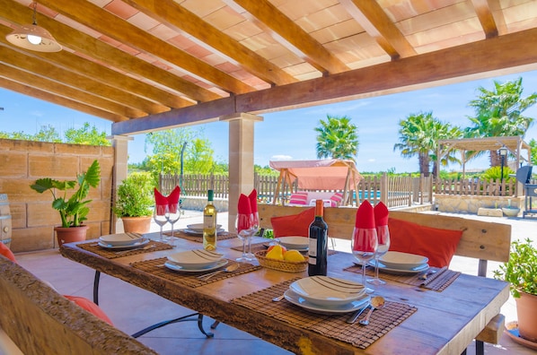Un área de comedor al aire libre con una pérgola de madera, mesas dispuestas con platos, cubiertos y copas de vino, rodeada de plantas en macetas y vistas a palmeras.