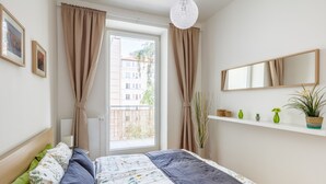 A bedroom with a bed, a window with curtains, a bedside lamp, a shelf with decorative items, and a mirror on the wall. - Beautiful Apartment with Private Terrace in Central Prague (Praha 3)