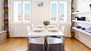 A dining area with a table set for four, two windows with curtains, a radiator, and a shelf with books and decorative items.