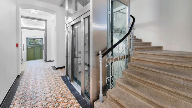 A hallway with patterned flooring, a staircase with a metal railing, and a door with a green sign.