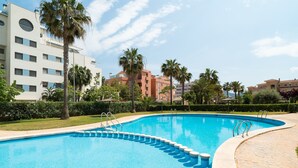 Une piscine à bord incurvé, entourée de palmiers et bordée d’un immeuble à plusieurs étages doté de balcons.