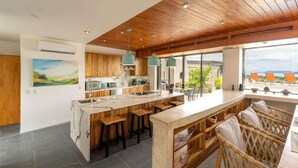 A modern kitchen with wooden cabinets, a large island with stools, pendant lights, and a view of the outdoors.