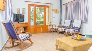 A room with a wooden desk, a television, a rocking chair, two lounge chairs, a wicker table with fruit, and a view of greenery through the door. - PENYA - Chalet with private garden in S'Illot. Free WiFi (Manacor)