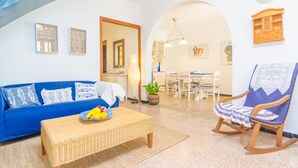 A living room with a blue sofa, wicker coffee table, and a rocking chair. There is a dining area in the background with a table and chairs. - PENYA - Chalet with private garden in S'Illot. Free WiFi (Manacor)
