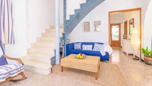 A living room with a blue sofa, a wicker coffee table, a rocking chair, a staircase, and a kitchen visible through an open doorway. - PENYA - Chalet with private garden in S'Illot. Free WiFi (Manacor)