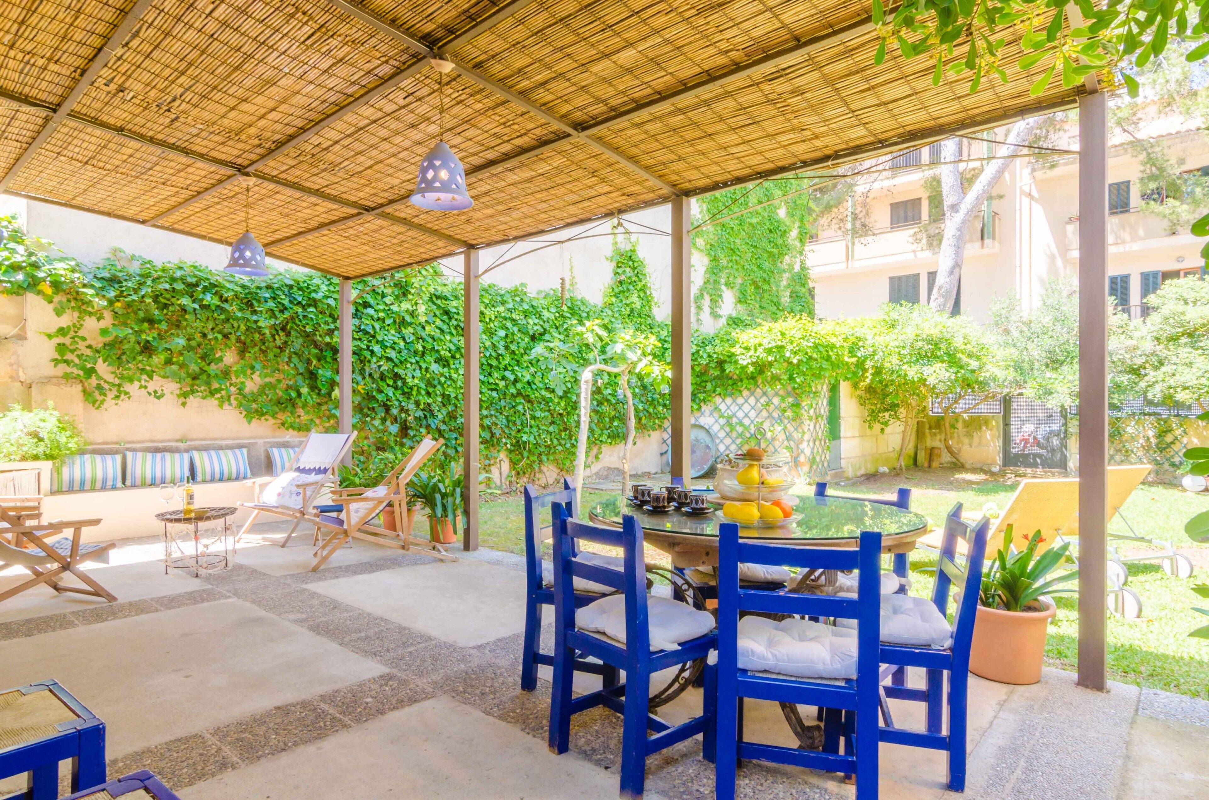 A covered outdoor seating area with blue chairs and a table, surrounded by greenery and a view of buildings.