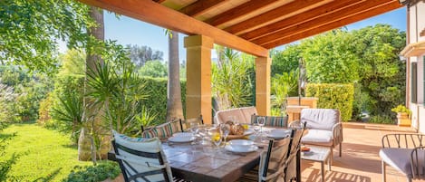 An outdoor patio with a wooden pergola, a dining table set with plates, glasses, and a bowl, surrounded by cushioned chairs.