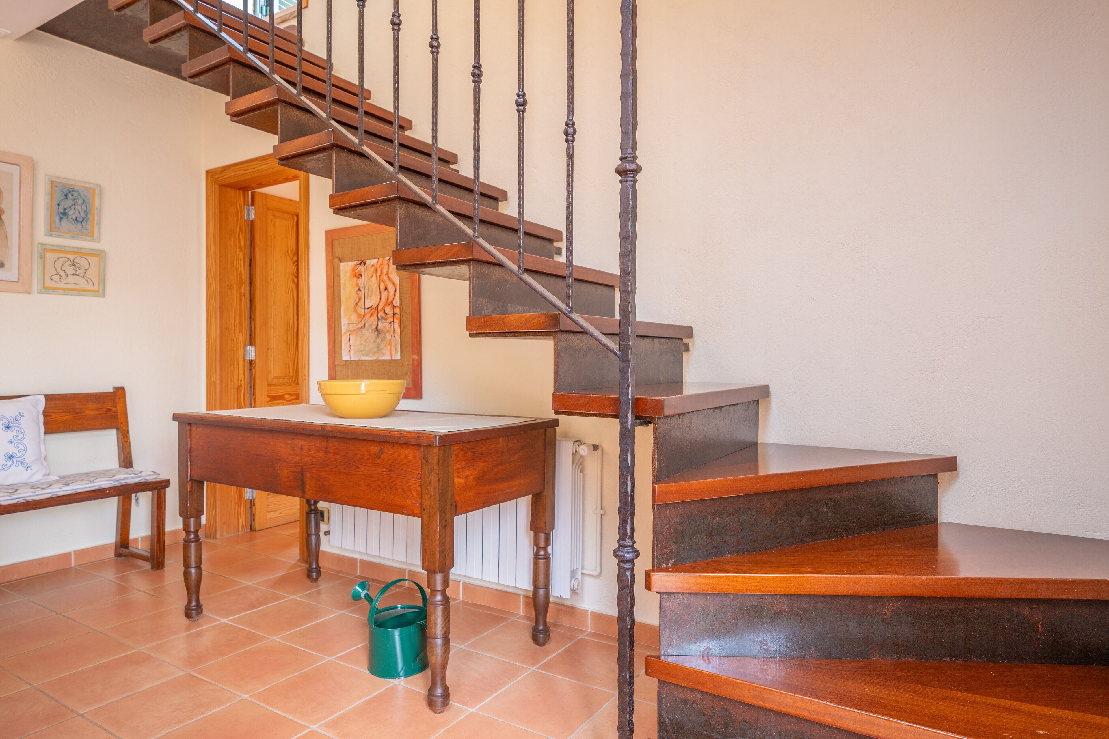 Un escalier en bois avec une rampe en métal, une table en bois sur laquelle se trouve un bol jaune, un seau vert et un banc.