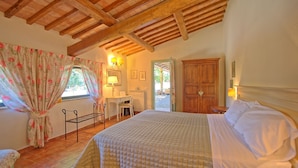 Quarto com teto de madeira, cama com colcha xadrez, escrivaninha com cadeira e janela com cortinas florais.