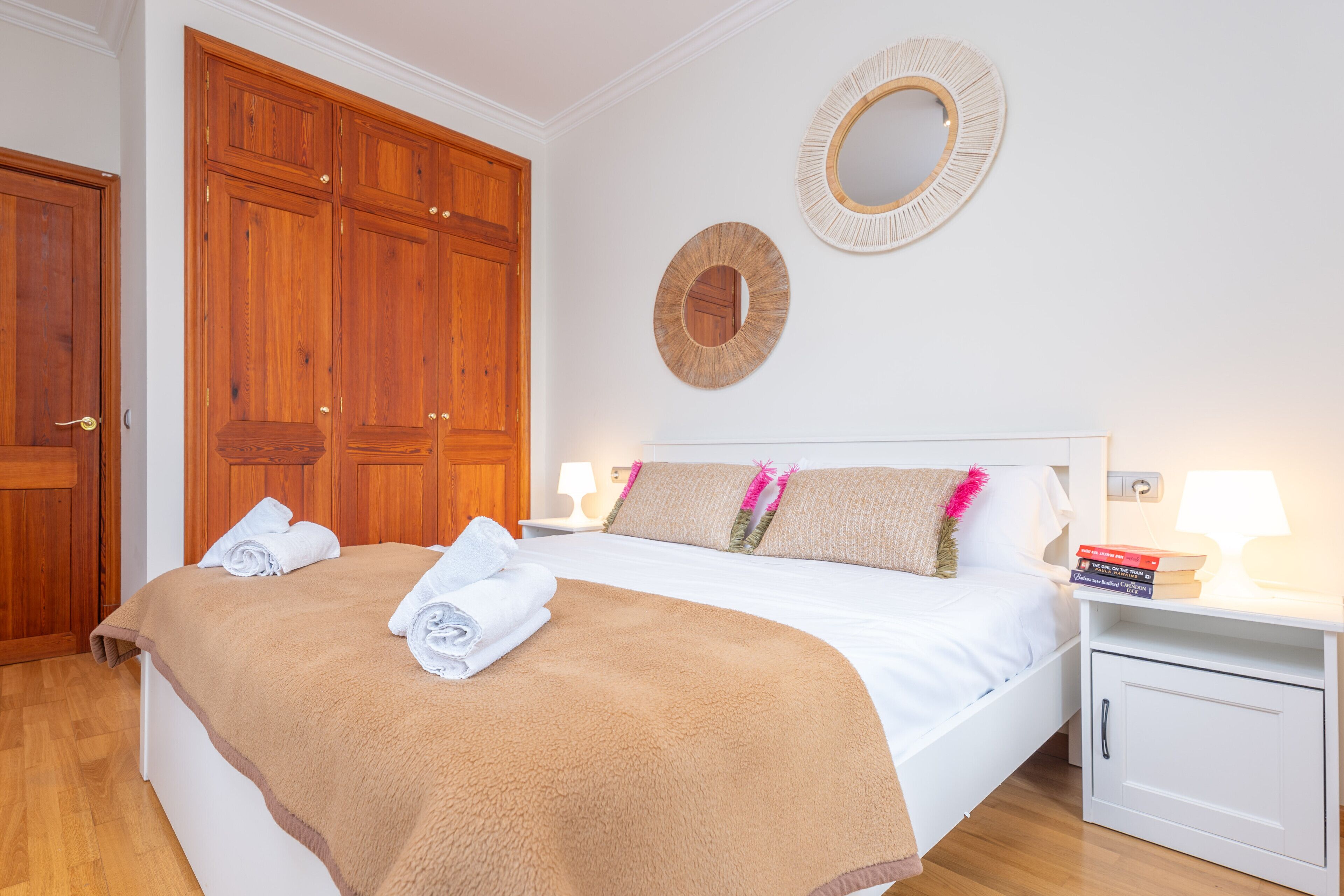 A neatly made bed with a brown blanket and rolled towels, wooden wardrobe, two wall mirrors, and bedside lamps.