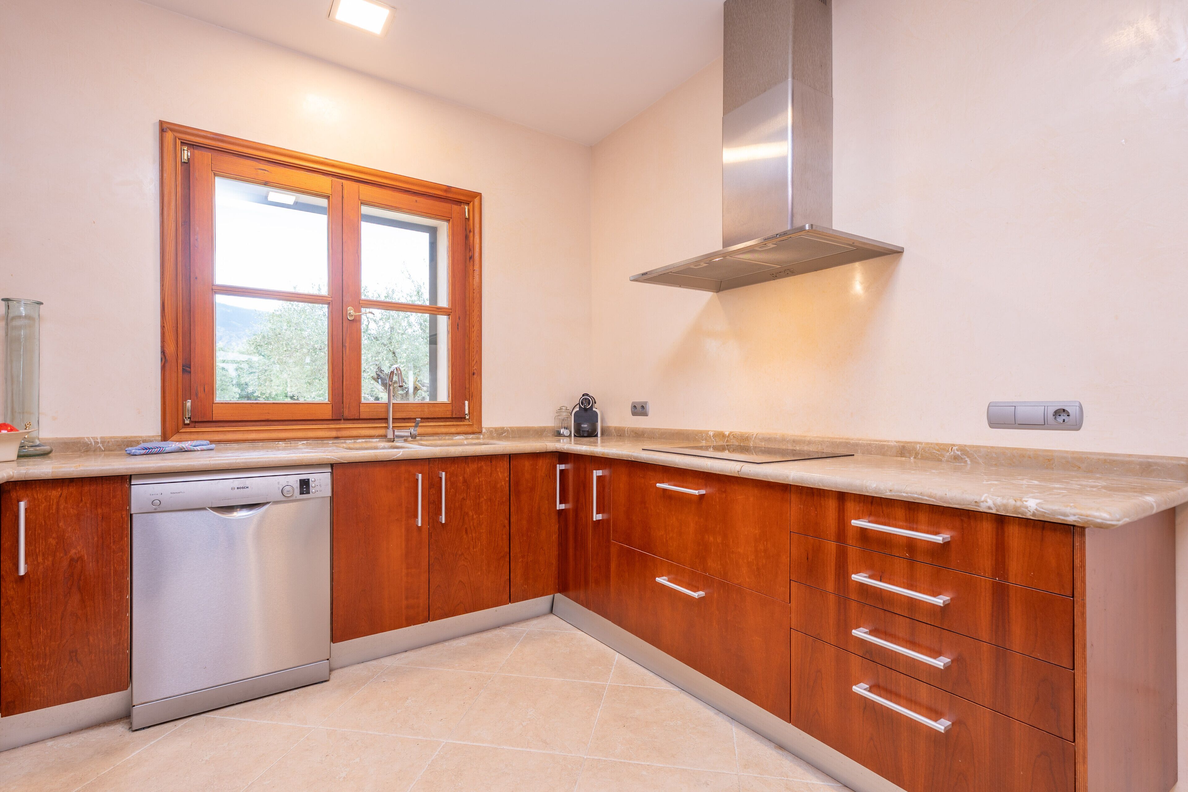 Una cocina con armarios de madera, lavavajillas, ventana y campana extractora de acero inoxidable.