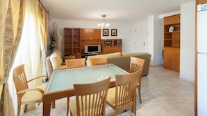 A living room with a glass-top dining table, wooden chairs, a sofa, a TV, and built-in wooden shelving. - CASCADES - Apartment with shared pool in Dénia. (Denia)