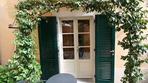 Un umbral con contraventanas verdes, un cristal y una pequeña mesa con una silla al frente.