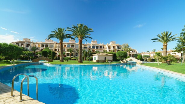 A swimming pool with a clear blue water, surrounded by palm trees and a building with multiple balconies in the background.