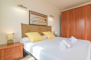 Una cama bien hecha con sábanas blancas y dos almohadas amarillas, una mesita de noche de madera con una lámpara y un armario de madera.
