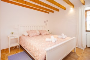 A bedroom with a bed, bedside tables, a nightstand, a window with curtains, and wooden beams on the ceiling.