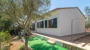 A white building with green shutters, a stone retaining wall, and a green artificial lawn. - ES JAÇ - Villa with terrace in Cala s'Almonia with free Wi-Fi (Cala Llombards)