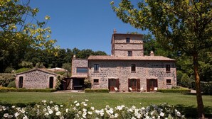 Un edificio de piedra con una torre central, una estructura más pequeña adosada y un jardín bien cuidado con flores blancas.