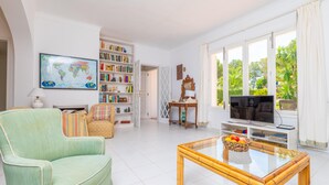 A living room with a sofa, a coffee table with fruit, a bookshelf, a TV, and a large window with a view of greenery. - CAN FERRO - Villa with private pool in Cala Blava. Free WiFi (Llucmajor)